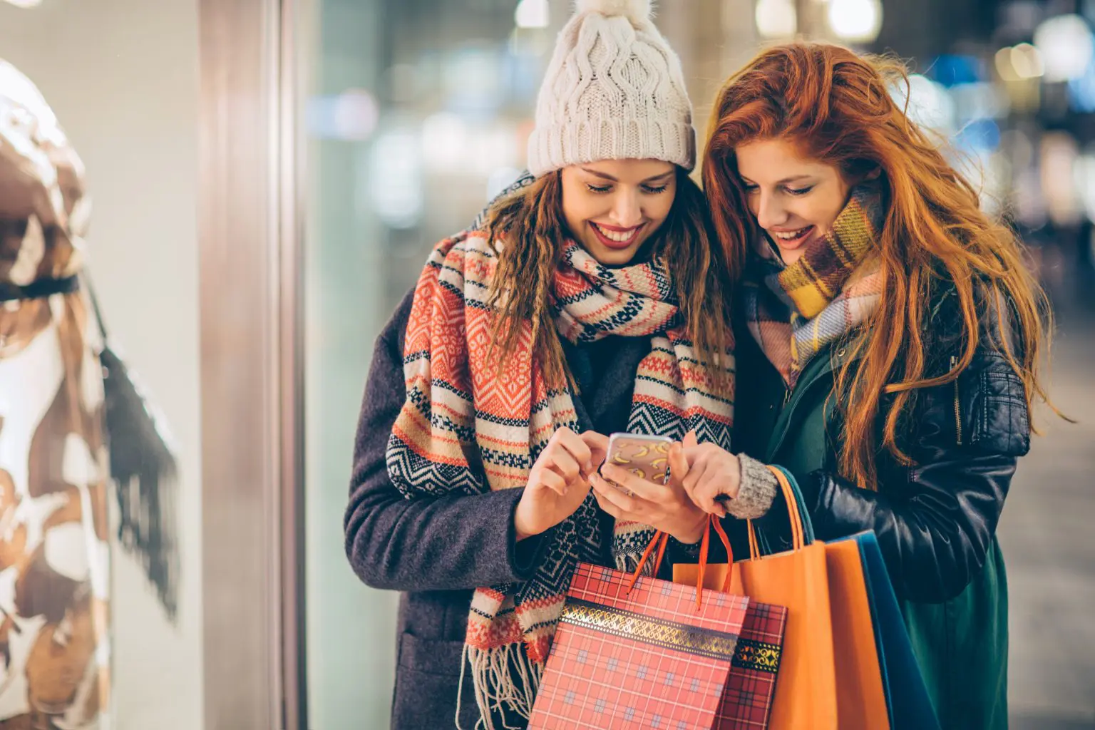 Friends walking on city street by night during Black Friday and Cyber Monday. Checking smart phone to see which shops they pick Wearing warm clothing.