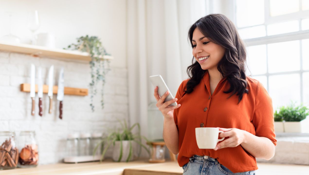 Woman standing in kitchen looking at phone and drinking coffee